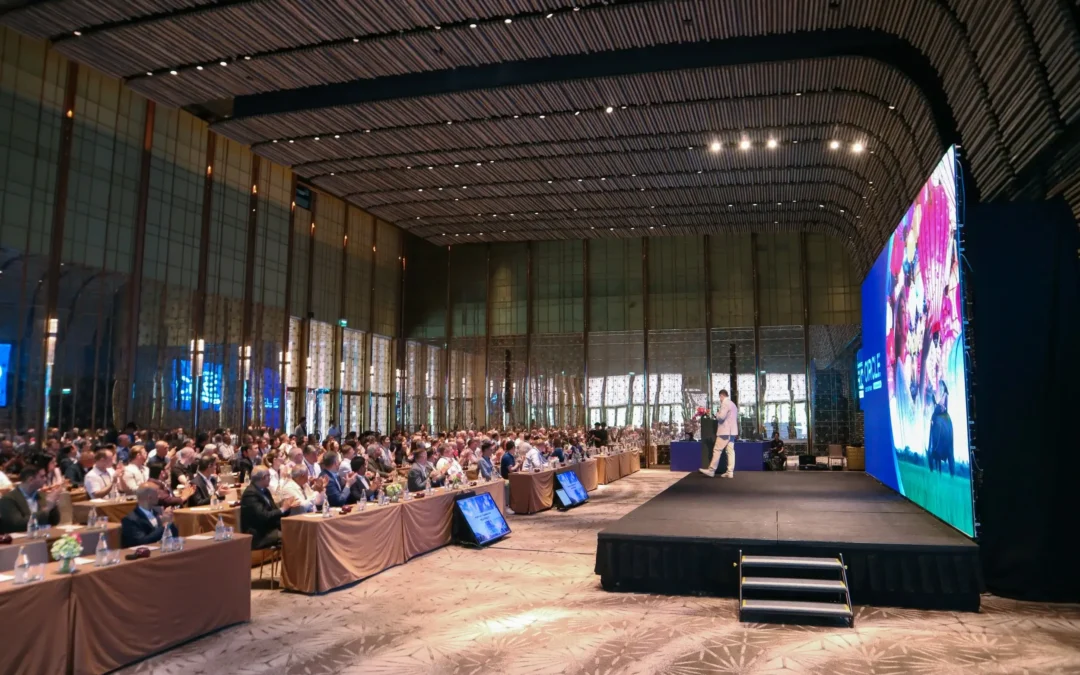 Speaker presenting on stage to a large seated audience in a modern conference hall with a large LED screen displaying colorful visuals.