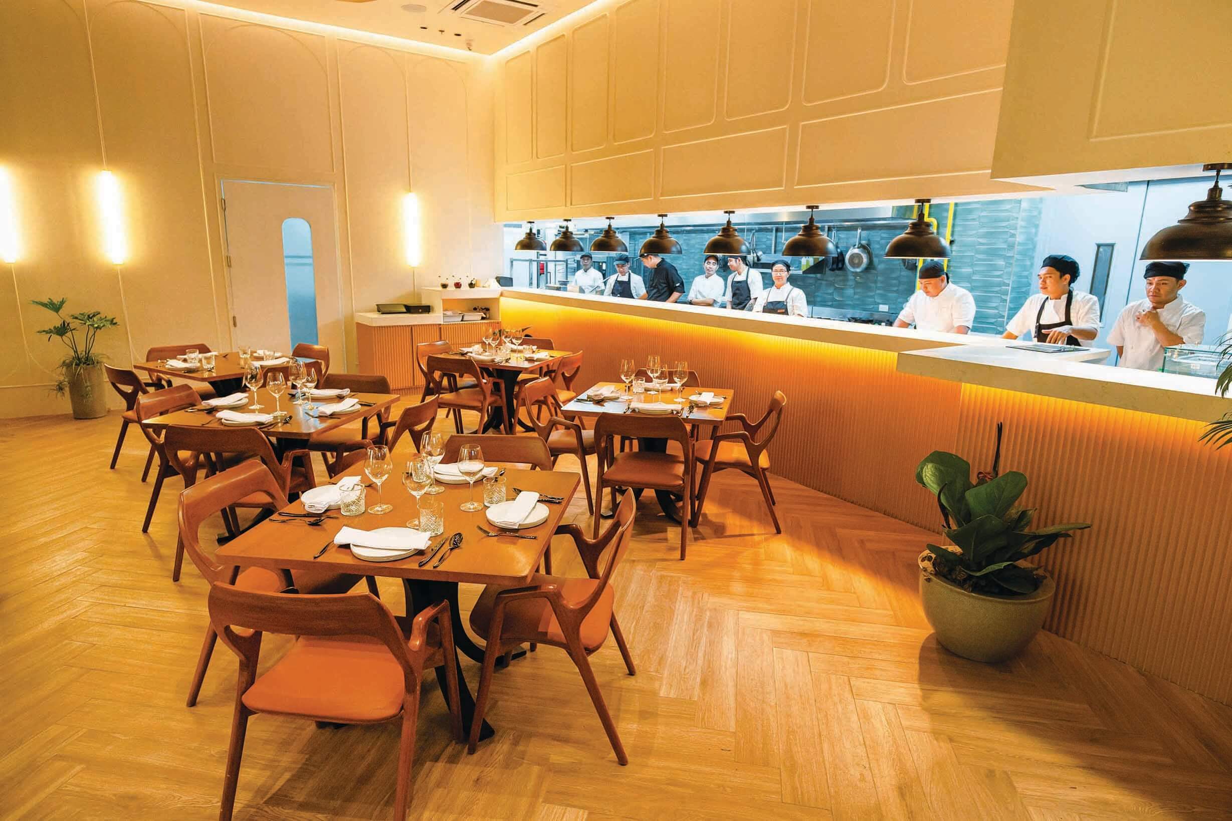 Modern open-kitchen restaurant interior with wooden flooring, mid-century style furniture, and chefs visible behind a service counter.
