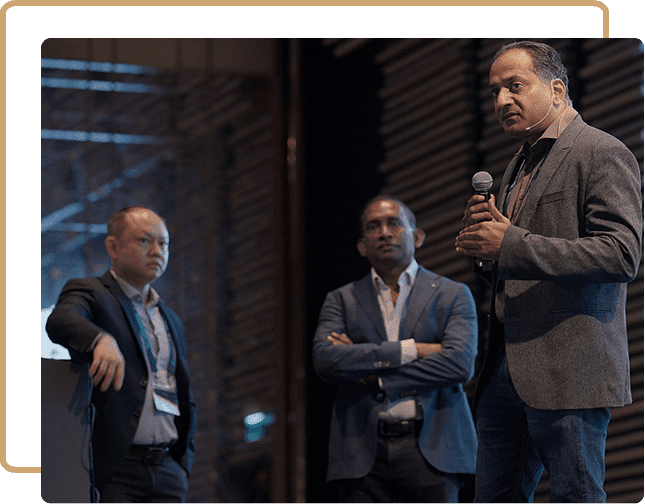 A group of three professionals engaged in a discussion or presentation at an event. One individual is speaking into a microphone, while the other two are standing with their arms crossed, listening attentively. They are dressed in business attire and appear to be on stage, likely during a conference or seminar. The setting is modern and sophisticated, with architectural elements visible in the background.