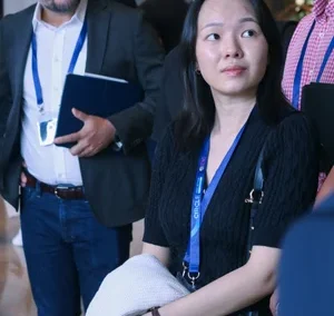 Woman with a conference badge stands holding a coat, surrounded by other professionally dressed attendees during a networking event.