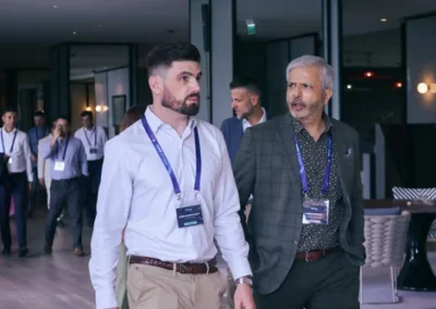 Two professionally dressed men wearing conference badges walk and converse in a modern venue, with other attendees in the background.
