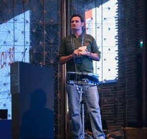 Man standing on a stage giving a presentation with a microphone headset and a small tech device on a stand beside him.