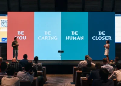 Two presenters on stage in front of a large screen displaying the words "Be You," "Be Caring," "Be Human," and "Be Closer" in colorful panels during a conference session.