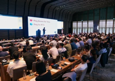 Large audience seated in a conference hall watching two presenters speak on stage in front of a screen displaying key messages.