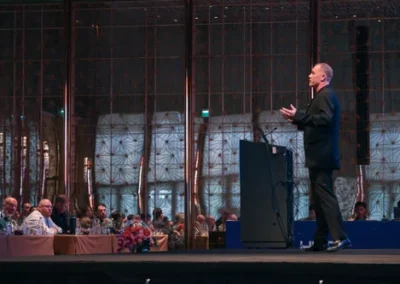 Male speaker presenting on stage at a formal conference, with seated attendees watching in a large, elegantly lit hall.