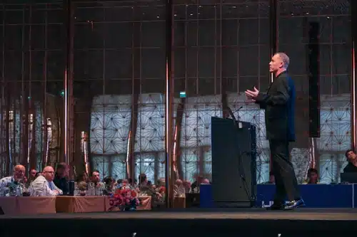 Male speaker presenting on stage at a formal conference, with seated attendees watching in a large, elegantly lit hall.