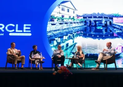 Five panelists seated on stage during a conference discussion, with a large screen behind them displaying a scenic riverside view.