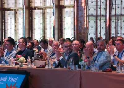 Audience of professionally dressed attendees clapping during a conference in a large, well-lit venue with floral table arrangements.
