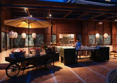 Outdoor refreshment station at night with a decorative cart, umbrella, and a bartender preparing drinks at a counter under warm lighting.
