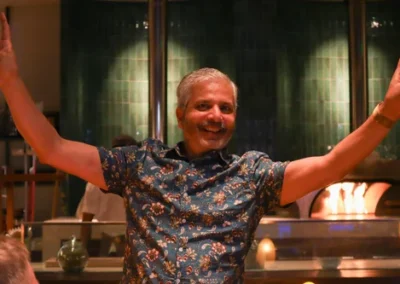 Smiling man in a floral shirt raising both arms joyfully in a warmly lit restaurant setting.