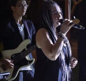 Female vocalist singing into a microphone during a nighttime outdoor performance with a guitarist in the background.