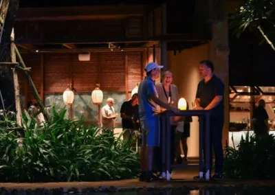 Group of people socializing around a tall table at an outdoor evening event near a restaurant with warm lantern lighting.