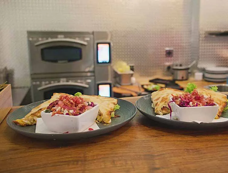 Two plated flatbread dishes on a counter in front of stacked rapid-cook ovens in a compact commercial kitchen