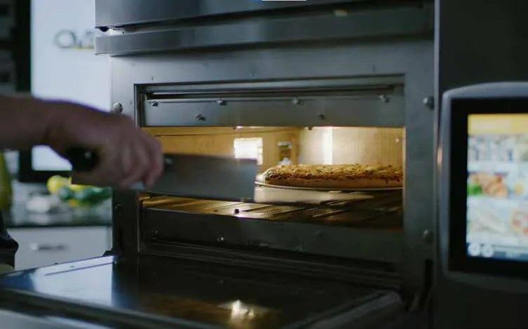 A chef sliding a pizza into a countertop rapid-cook oven using a peel